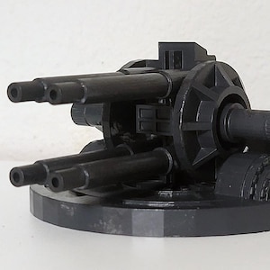Puede incluir: Un cañón de juguete gris oscuro con múltiples cañones y una base circular. El juguete presenta múltiples cañones y un diseño mecánico detallado, lo que sugiere un tema militar o de ciencia ficción en miniatura.