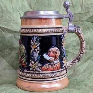 May include: A ceramic beer stein with a pewter lid and handle. The stein features a scene of a man and a dog in a woodland setting with flowers and leaves. The stein is decorated with a brown, black, green, and white colour scheme.