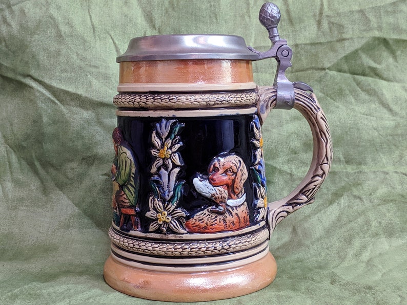 May include: A ceramic beer stein with a pewter lid and handle. The stein features a scene of a man and a dog in a woodland setting with flowers and leaves. The stein is decorated with a brown, black, green, and white colour scheme.