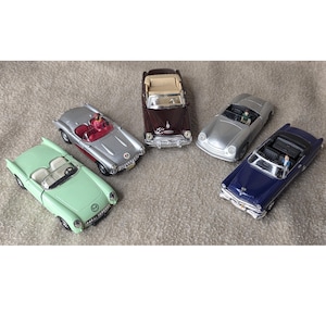May include: Five miniature classic convertible cars in various colors: mint green, silver, burgundy, silver, and navy blue. Each car has detailed features, including open tops and interior details. The cars are arranged on a neutral-colored surface.