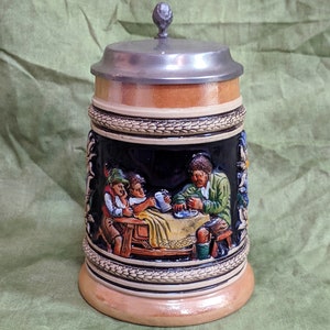 May include: A ceramic beer stein with a pewter lid. The stein features a relief scene of three men sitting at a table, drinking beer and eating. The scene is painted in bright colours, including green, blue, red, and yellow.