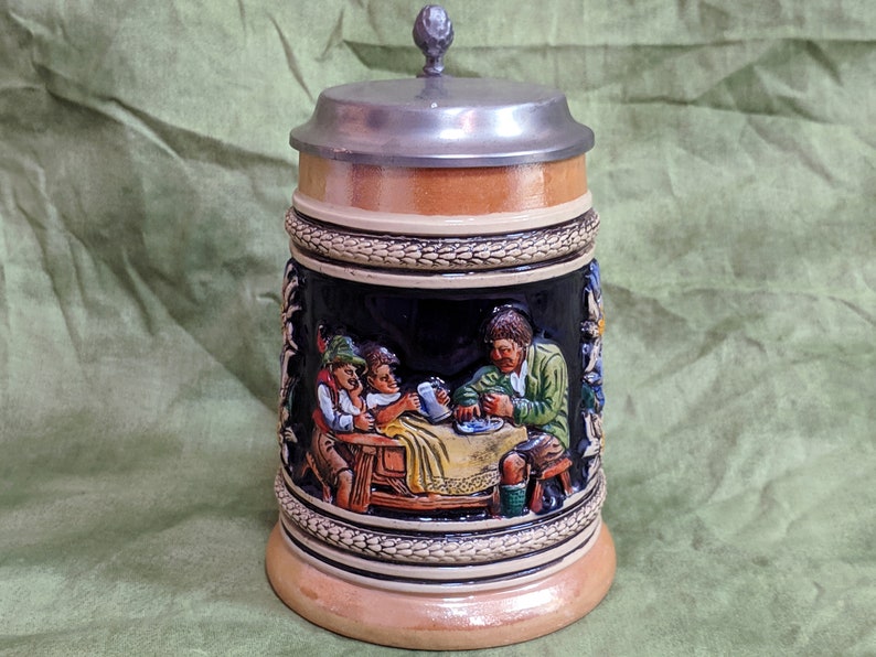 May include: A ceramic beer stein with a pewter lid. The stein features a relief scene of three men sitting at a table, drinking beer and eating. The scene is painted in bright colours, including green, blue, red, and yellow.