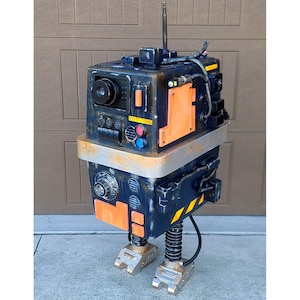 以下が含まれることがあります： 銀色のアンテナ、丸い黒い目、銀色の台座が付いた黒とオレンジのロボット。ロボットの正面には、「注意：高電圧」と書かれた黄色の警告表示があります。