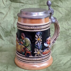 May include: A ceramic beer stein with a pewter lid and handle. The stein features a relief design of people and flowers.