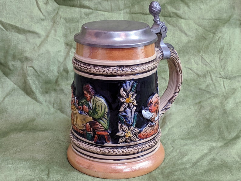 May include: A ceramic beer stein with a pewter lid and handle. The stein features a relief design of people and flowers.