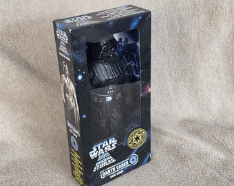 May include: A Star Wars Darth Vader action figure in a black and blue box. The box features the Star Wars logo, character images, and the text "Darth Vader". The figure is visible through a clear window.