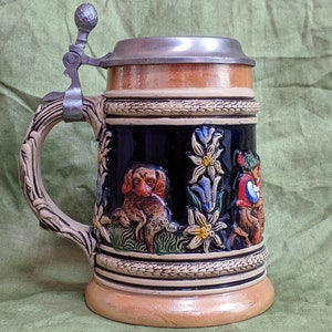 May include: A brown and white ceramic beer stein with a pewter lid. The stein features a detailed design of a dog, flowers, and a person in traditional clothing.