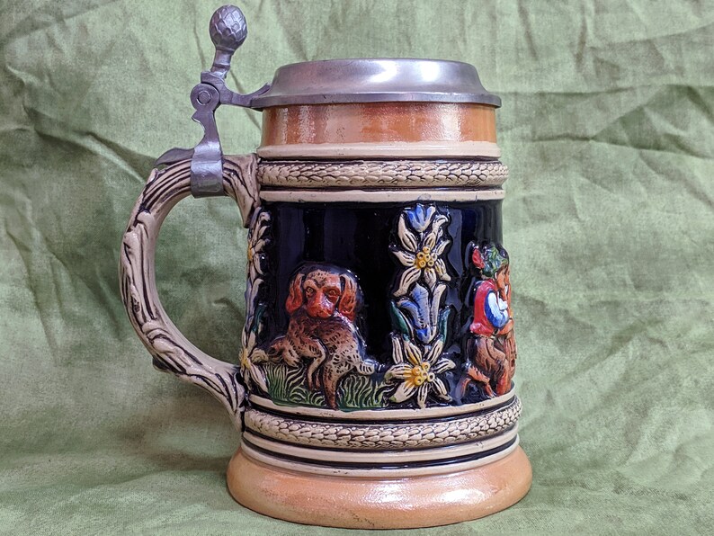 May include: A brown and white ceramic beer stein with a pewter lid. The stein features a detailed design of a dog, flowers, and a person in traditional clothing.