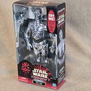 May include: A silver TC-14 action figure from Star Wars Episode I, displayed in its original packaging. The box features the Star Wars logo, the text "TRY ME!" and "ELECTRONIC TC-14". The figure is visible through a clear plastic window.
