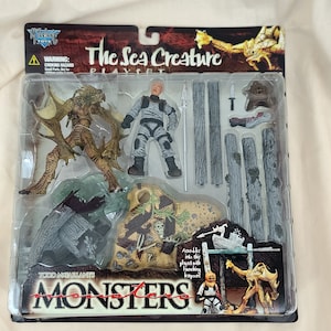 May include: A packaged "The Sea Creature" playset featuring monster figures, a human figure, and accessories. The package includes the text "Monsters" and "Todd McFarlane Toys". The figures are in shades of brown, tan, and gray.