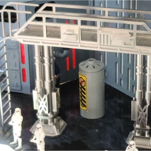 Puede incluir: Un juego de Star Wars con una estructura de pórtico blanca sostenida por columnas plateadas. Se ve un contenedor cilíndrico gris con una etiqueta amarilla y negra. Figuras de Stormtroopers están en el suelo.