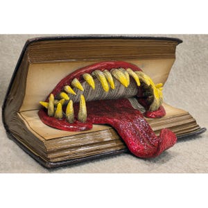 May include: A sculpted open book with a monster mouth emerging from the pages. The mouth is red with yellow teeth and a long red tongue. The book has aged, yellowed pages and a dark brown cover.