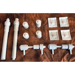 May include: A collection of white plastic model parts, including long cylindrical pieces, small cylindrical components, and rectangular pieces with circular details. The parts are arranged on a brown surface.