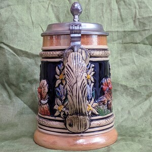 May include: A ceramic beer stein with a pewter lid and a wooden base. The stein features a detailed design of a traditional German scene with flowers, leaves, and figures. The handle is shaped like a tree trunk.