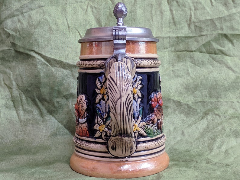May include: A ceramic beer stein with a pewter lid and a wooden base. The stein features a detailed design of a traditional German scene with flowers, leaves, and figures. The handle is shaped like a tree trunk.