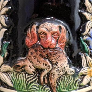 May include: A ceramic beer stein featuring a detailed relief design of a brown and red dog holding a smaller brown and white dog in its paws. The dog is surrounded by green grass and white flowers with yellow centres.