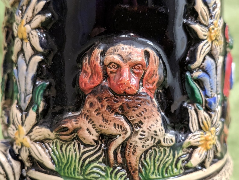 May include: A ceramic beer stein featuring a detailed relief design of a brown and red dog holding a smaller brown and white dog in its paws. The dog is surrounded by green grass and white flowers with yellow centres.