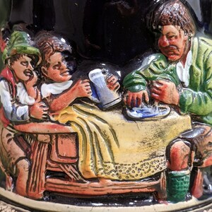 May include: A ceramic stein featuring a scene of three people enjoying a beer. The stein is decorated with a dark blue glaze and depicts a man, a woman, and a child seated at a table. The man is wearing a green jacket and has a moustache. The woman is wearing a yellow dress and has long brown hair. The child is wearing a red and white shirt and has short brown hair. The stein is a traditional German design.