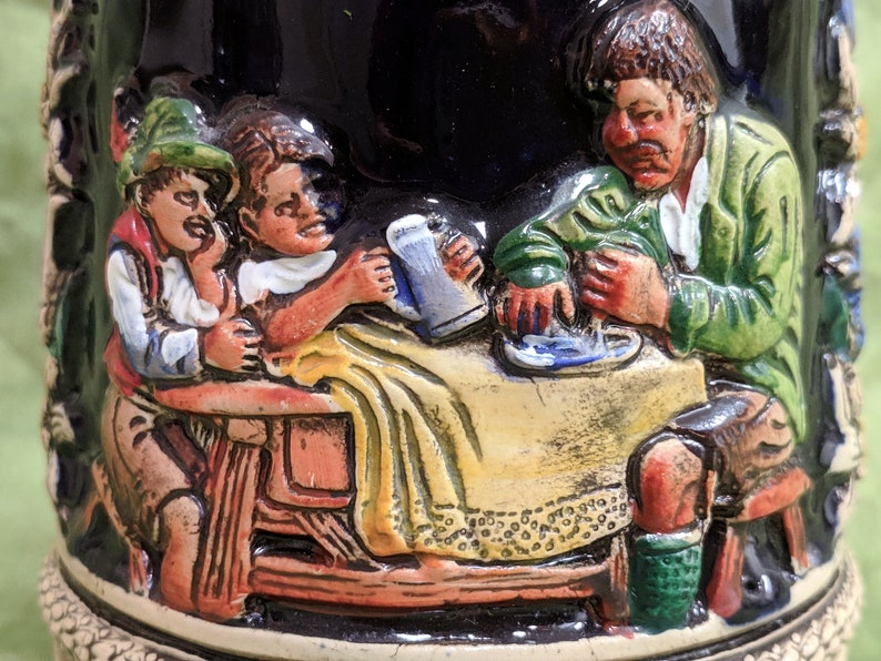 May include: A ceramic stein featuring a scene of three people enjoying a beer. The stein is decorated with a dark blue glaze and depicts a man, a woman, and a child seated at a table. The man is wearing a green jacket and has a moustache. The woman is wearing a yellow dress and has long brown hair. The child is wearing a red and white shirt and has short brown hair. The stein is a traditional German design.