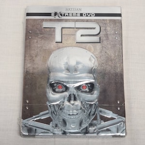 May include: A silver DVD case with a metallic background and the text "Artisan Home Entertainment Extreme DVD T2" on the front. A close-up of a robotic face with red eyes is featured in the center.