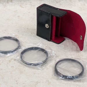 May include: Three round, clear glass camera filters in individual plastic bags, with a black leather case with a red lining.