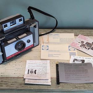 May include: A vintage Polaroid Land Camera Automatic 250 with a black and silver body, a red button, and a black strap. The camera is surrounded by instruction manuals and a pamphlet with the text "See your dealer or use this order mailer" and "Order prints, copy prints and enlargements".
