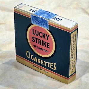 May include: A vintage Lucky Strike cigarette pack. The pack is dark blue with a red and yellow circular logo that reads "LUCKY STRIKE" and "IT'S TOASTED". The text "CIGARETTES" and "MADE IN USA" are also visible.