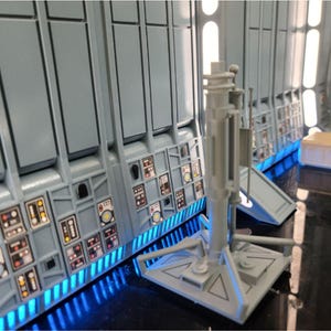 May include: A detailed model of a spaceship interior, featuring gray wall panels with control panels and blue LED lighting. A central support structure stands in the foreground, with a small yellow container visible in the background. The scene evokes a futuristic or science fiction theme.