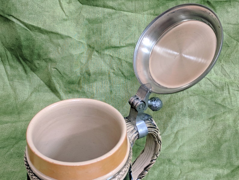 May include: A ceramic beer stein with a hinged lid. The stein has a brown and white design and a green base. The lid is made of metal and has a shiny finish.