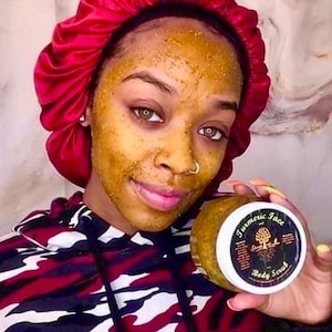May include: A person wearing a red satin bonnet and a red, black, and white camouflage hoodie, with a yellow-toned granular scrub applied to their face. They are holding a clear plastic jar of Turmeric Face and Body Scrub with a white lid.