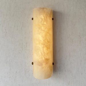 May include: A vertical, semi-cylindrical wall sconce featuring a translucent, cream-colored shade with a marbled, cloud-like texture. The shade is secured to the wall by four small, circular, dark metallic fasteners positioned on the sides.