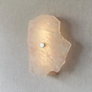 May include: A wall-mounted light fixture made from a translucent, irregularly shaped stone. The stone has a soft pink hue with internal veining and a small, round, silver-colored button in the center. The light glows from within.