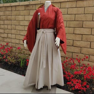May include: A traditional Japanese outfit featuring a red kimono and a white hakama. The kimono has long sleeves and a wide collar. The hakama is a pleated skirt worn over the kimono. The outfit is being worn by a person standing in front of a brick wall.