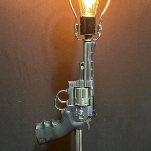 May include: A table lamp featuring a silver-toned revolver base with a black grip, mounted on a circular black stand. The lamp includes a vertical metal harp supporting a single glowing Edison-style light bulb that emits a warm yellow light.
