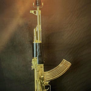 May include: A gold-colored rifle-shaped lamp featuring a black handguard and pistol grip. The barrel is topped with a glowing Edison-style light bulb. The structure is mounted on a circular black base, creating a vertical lighting fixture.