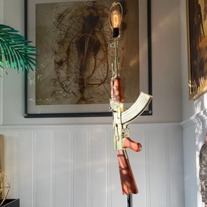 May include: A floor lamp featuring a gold-toned AK-47 rifle replica as the central stand, with a light bulb socket mounted at the top of the barrel. The rifle includes brown wood-grain textured furniture on the stock and pistol grip.