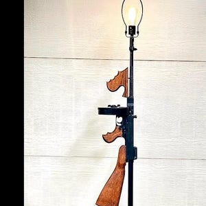 May include: A floor lamp featuring a black metal stand and a circular base. The central column incorporates a replica Thompson submachine gun with a dark wood stock, grip, and foregrip. A single light bulb is mounted at the top with a wire harp.