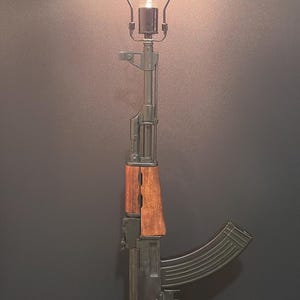 May include: A table lamp constructed from a rifle replica featuring a dark metal barrel and receiver, a wooden handguard and pistol grip, and a curved magazine. A metal harp supports a single glowing light bulb at the top of the rifle barrel.