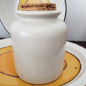 May include: A cream-colored ceramic jar with a cork stopper. The jar has a cylindrical shape with a slightly flared rim. The cork stopper is a light brown color. The jar is sitting on a decorative plate with a yellow and brown design.