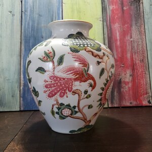 May include: White ceramic vase with a floral and bird design. The vase has a wide mouth and a round body. The design features pink and red birds with green and gold leaves and flowers.