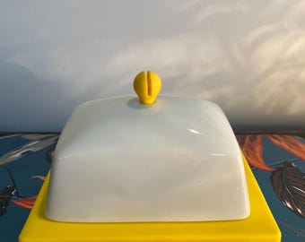 May include: A butter dish with a white lid and a bright yellow base. The lid has a small, yellow handle on top. The dish is rectangular and sits on a yellow base. The overall design is simple and modern.