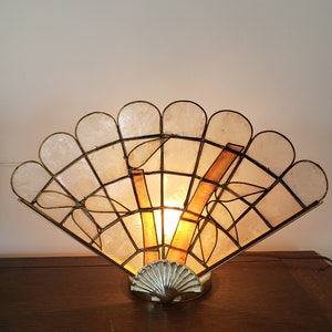 Peut inclure: Une lampe de table décorative avec un abat-jour en forme d'éventail en matériau blanc translucide. L'abat-jour est encadré d'une structure métallique et présente une base en forme de coquille.