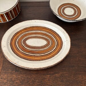 May include: A set of three white ceramic bowls with brown and tan concentric circle patterns. The bowls are on a dark wood surface.
