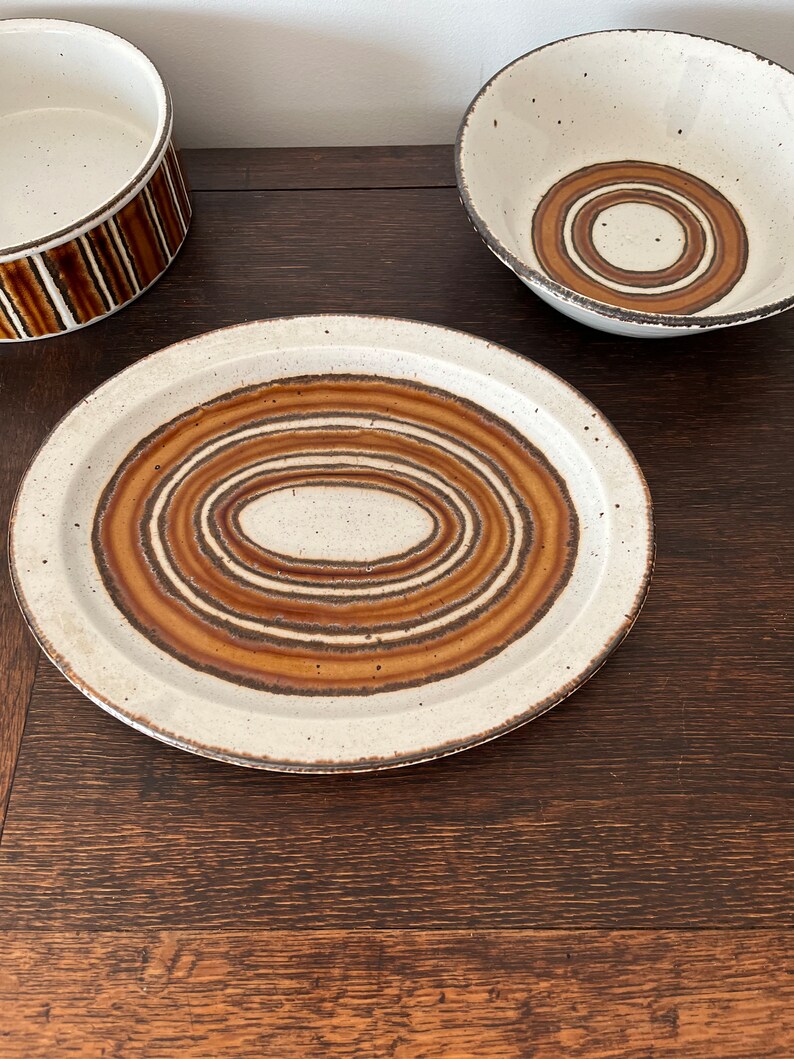 May include: A set of three white ceramic bowls with brown and tan concentric circle patterns. The bowls are on a dark wood surface.