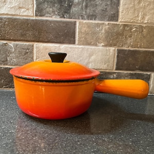 May include: An orange cast iron casserole pot with a lid and handle. The pot's colour graduates from red to orange. The lid has a black knob. The pot is approximately 20 cm in diameter, ideal for cooking stews and soups.