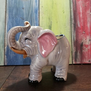May include: A grey ceramic elephant planter with a pink ear and a brown trunk. The planter has black feet and is sitting on a wooden surface.