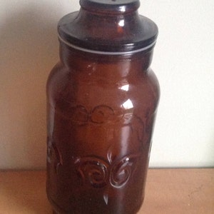 May include: A brown glass jar with a lid, featuring a decorative embossed design. The jar has a wide mouth and a rounded bottom.