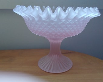 May include: A pink frosted glass pedestal bowl with a ruffled rim. The bowl has a diamond pattern on the body and a fluted base. The bowl is likely vintage and suitable for serving fruit or displaying decorative items.
