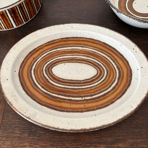 May include: A white ceramic plate with a brown and white concentric circle design. The plate has a slightly raised rim.
