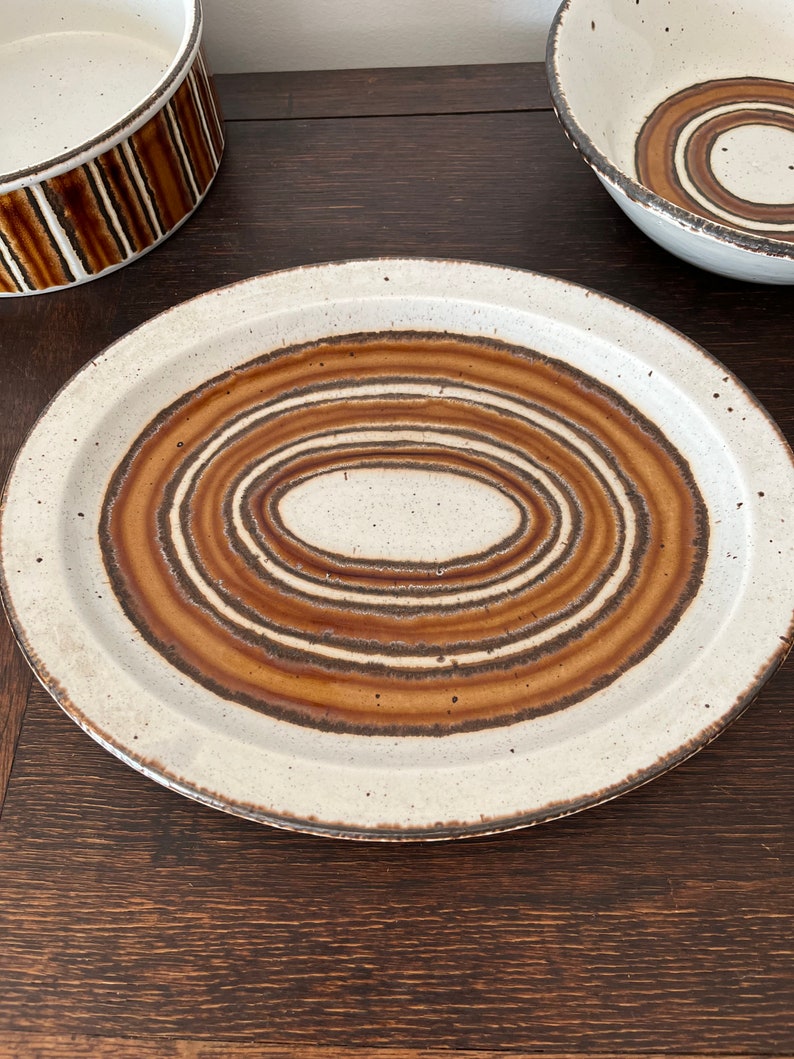 May include: A white ceramic plate with a brown and white concentric circle design. The plate has a slightly raised rim.